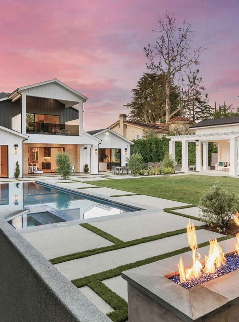 A modern backyard with a swimming pool, fire feature, outdoor seating, and a large house with balconies, all under a pink sunset sky.