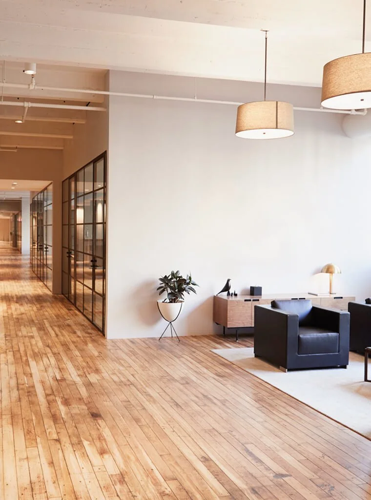 Modern office or lobby area with wooden flooring, glass-walled offices on the left, and a seating area with black armchairs, a wooden side table, a potted plant, and a table lamp on the right. Two large hanging ceiling lights are also visible.