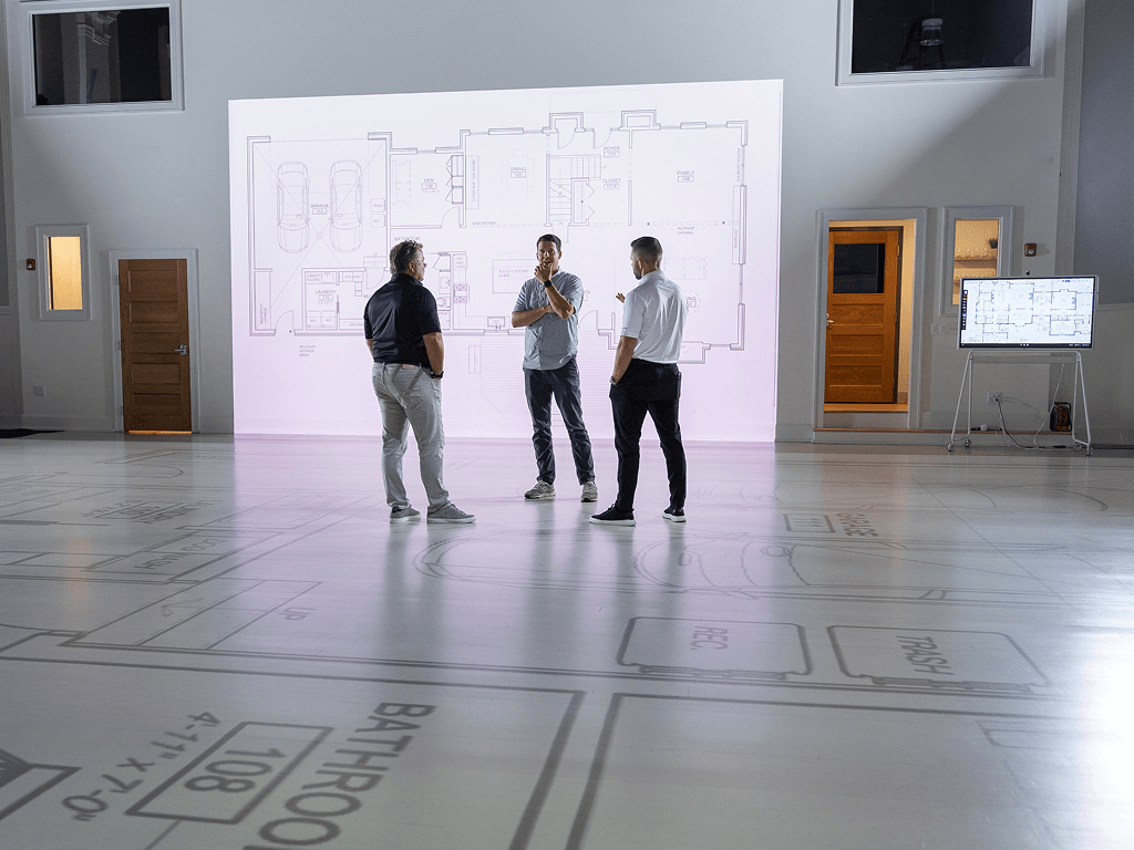 Three men are standing and discussing in front of a large illuminated architectural floor plan projection on a white wall in a spacious room with a glossy floor that has floor plans and measurements marked on it. There are two small windows and two doors on the wall.
