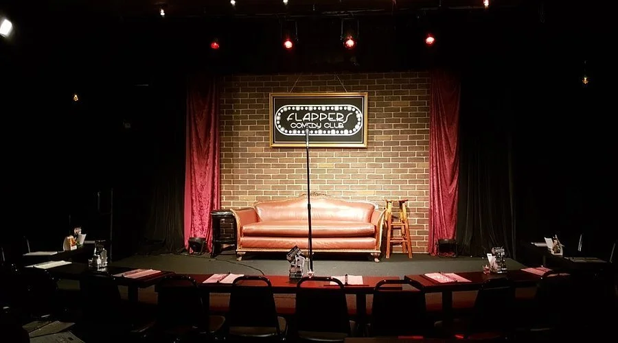 Empty comedy club stage with a sofa, a stool, and a brick wall backdrop, with red curtains on both sides and a sign that says 'The Flapper Comedy Club'.