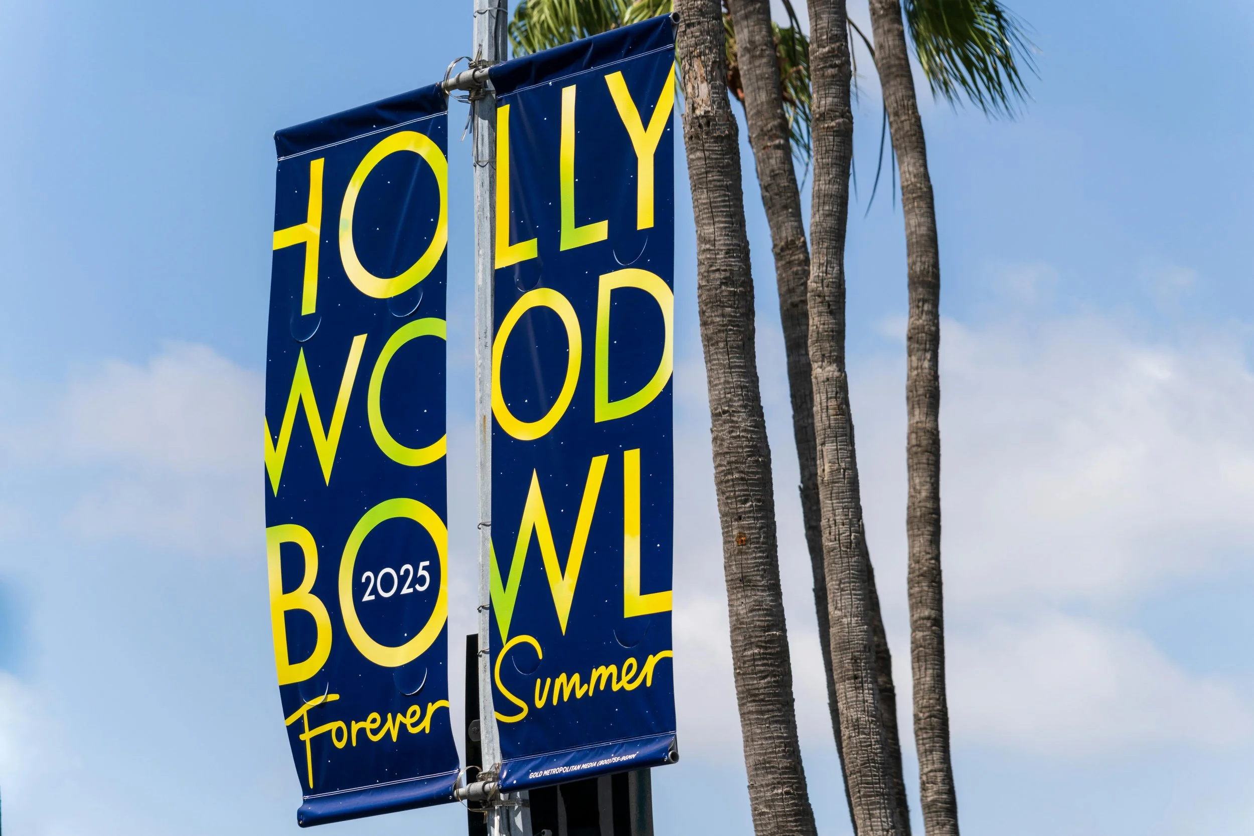 A sign with yellow and green text on a blue background that reads "Hollywood Bowl 2025 Forever Summer". The sign is attached to a pole next to three tall palm trees, under a blue sky with some clouds.