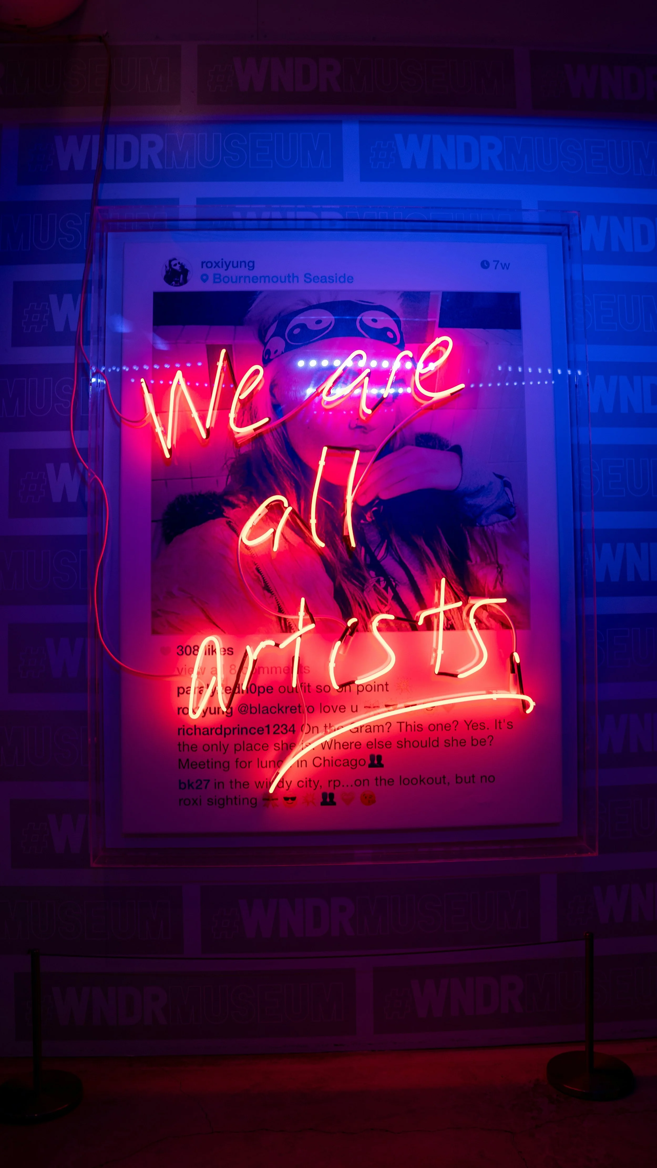Neon sign with pink and red lights spelling "we are artists" over a photograph of a woman wearing a bandana and jacket, displayed on a wall with posters in the background.