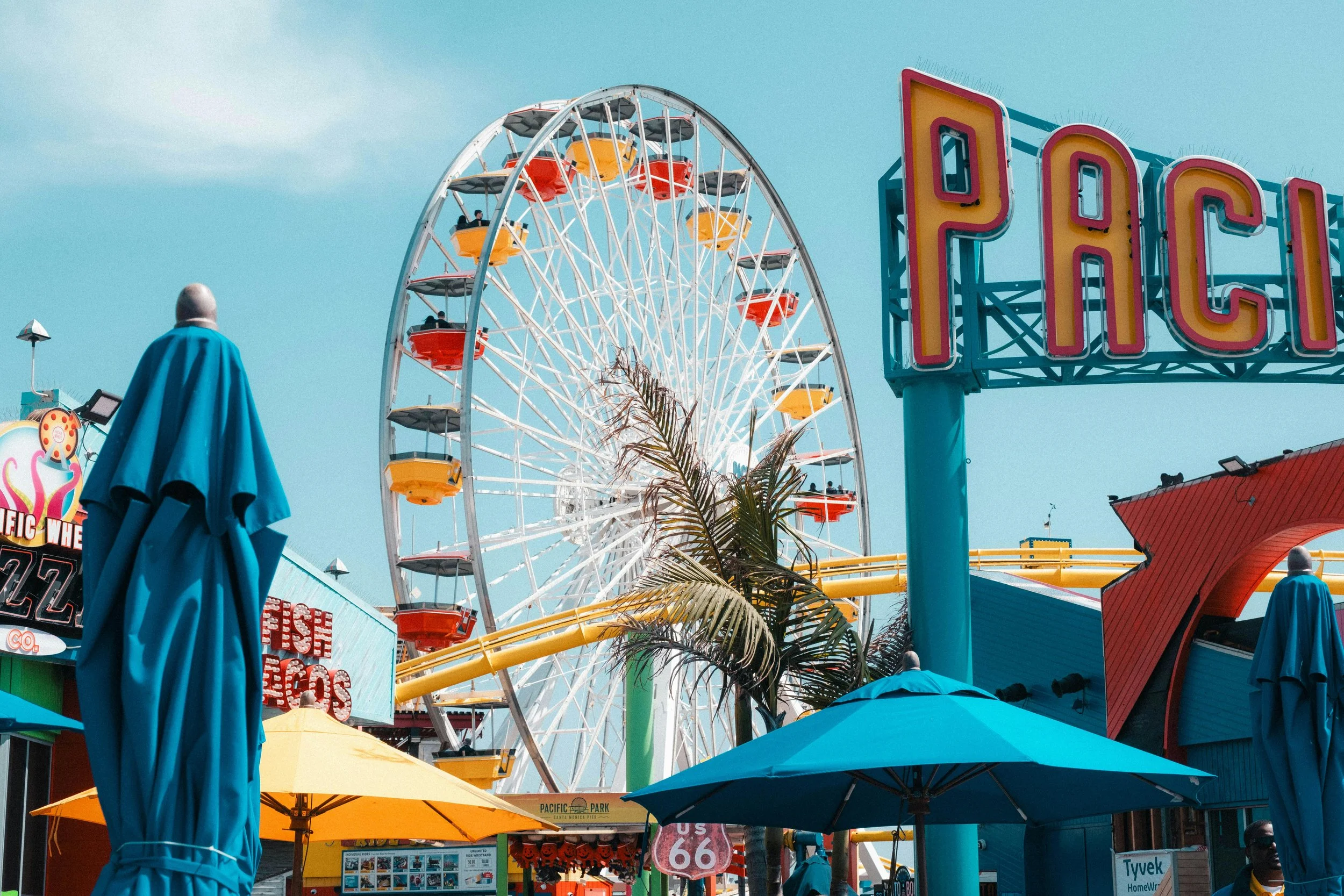 A large Ferris wheel with colorful seats at Pacific Park amusement park on a sunny day, with signs and umbrellas in the foreground.