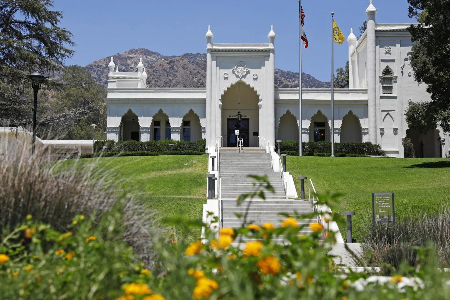 A white architectural building with Moorish design elements, stairs leading up to the entrance, surrounded by green grass and flowers, with flagpoles and mountain in the background.
