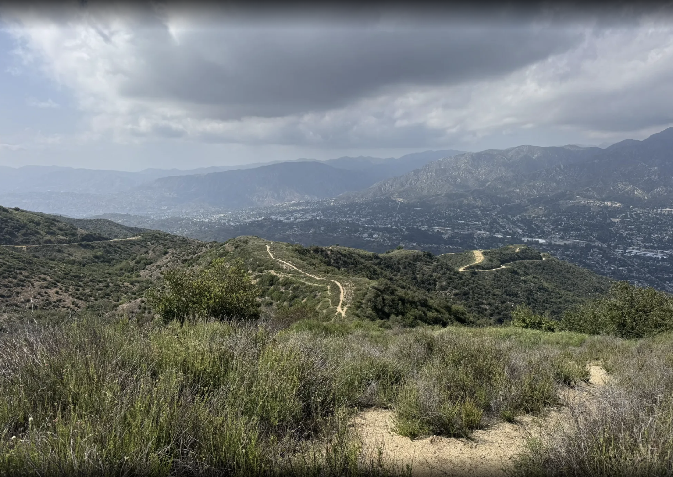 Hiking trail winding through green hills with mountains in the background under cloudy sky.