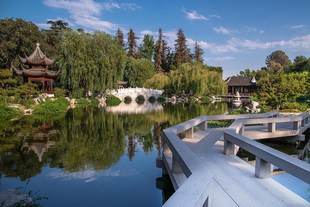 Serene Asian-style garden with traditional pagoda, arched stone bridge over pond, lush trees, and clear blue sky.