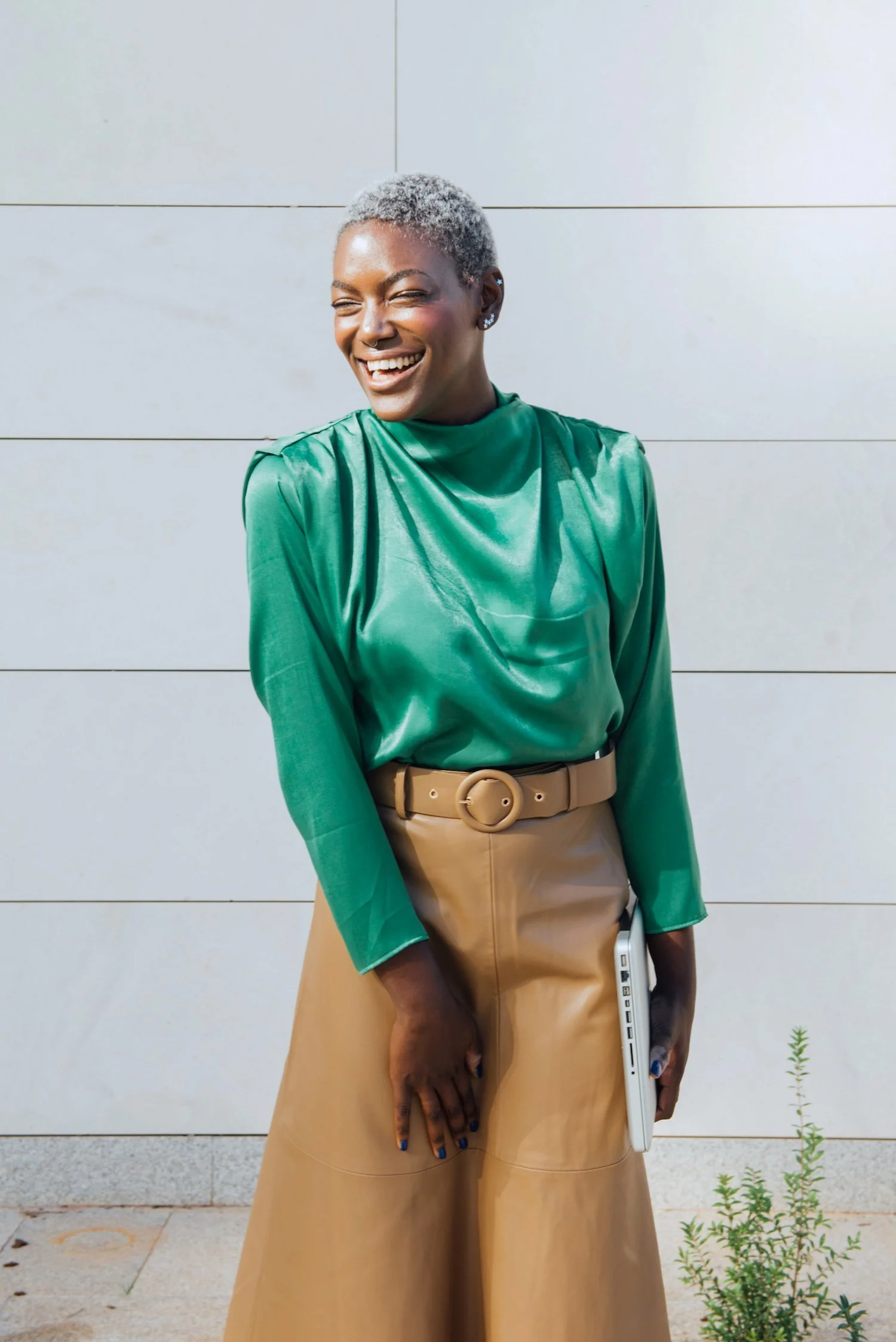 A woman with short gray hair and dark skin, wearing a shiny green top and tan pants with a matching belt, smiling and laughing while standing outdoors against a light-colored wall.