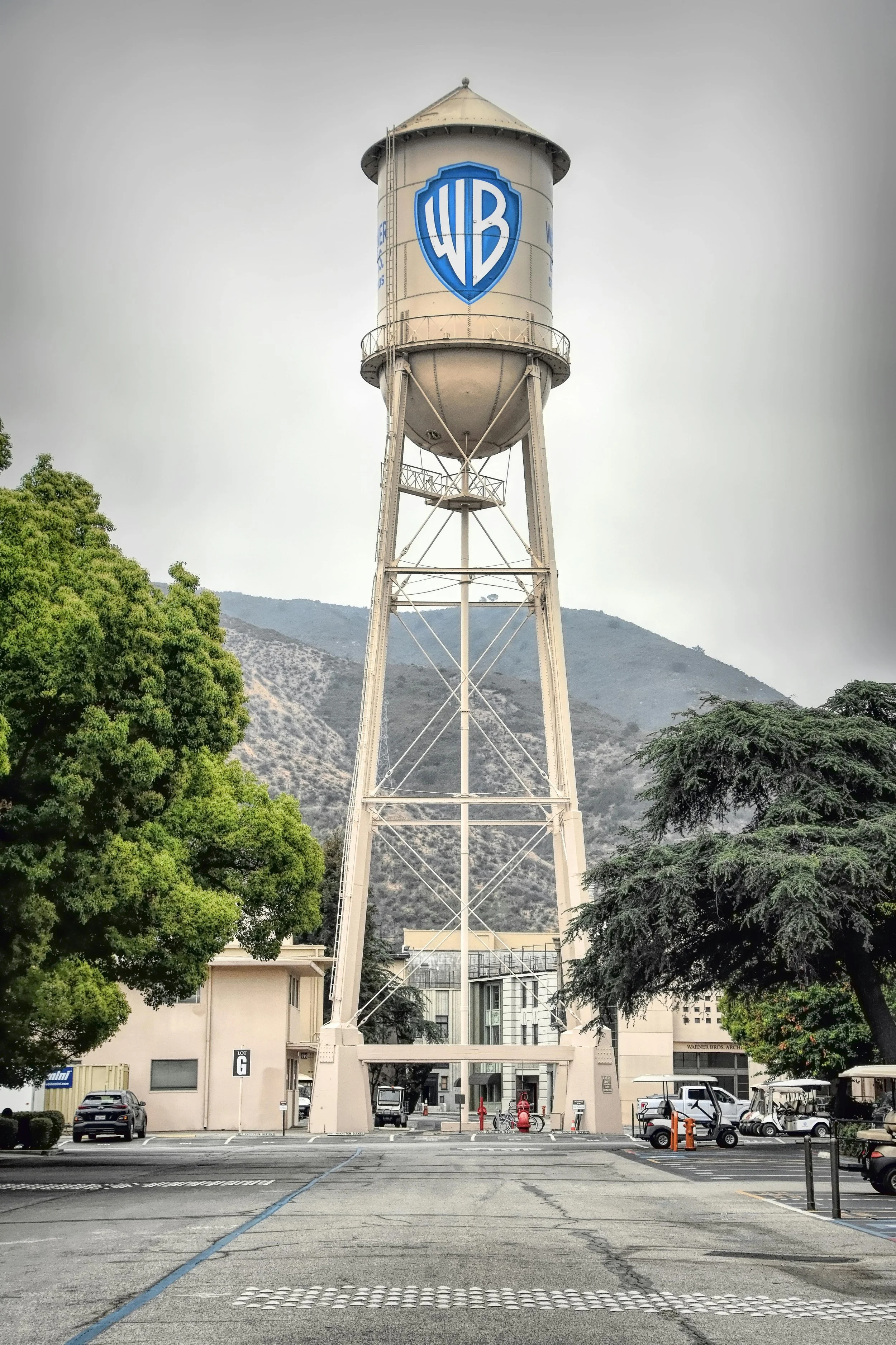 A tall water tower with the Warner Bros. logo on it, situated in an urban area with trees, cars, and mountains in the background.