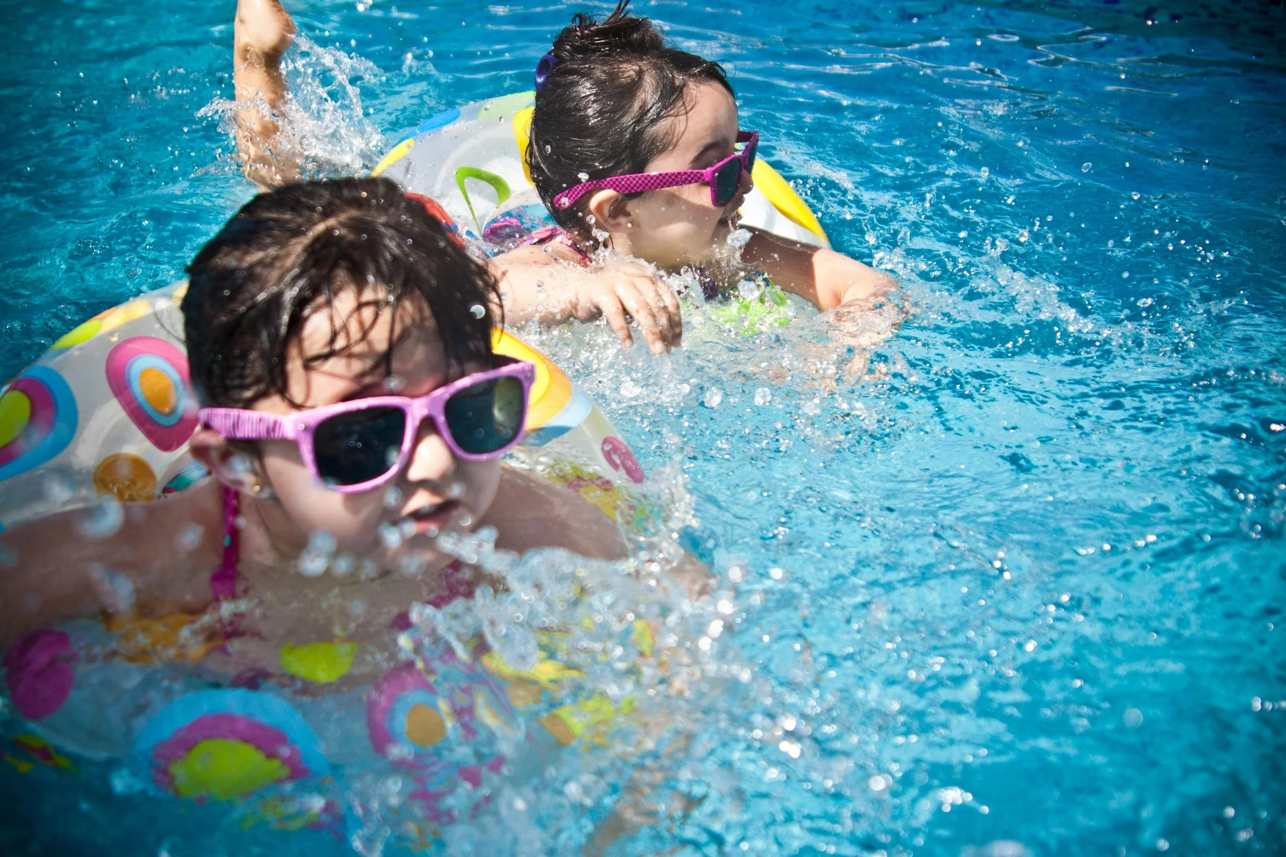Two young girls swimming in a pool with floatation rings wearing sunglasses.