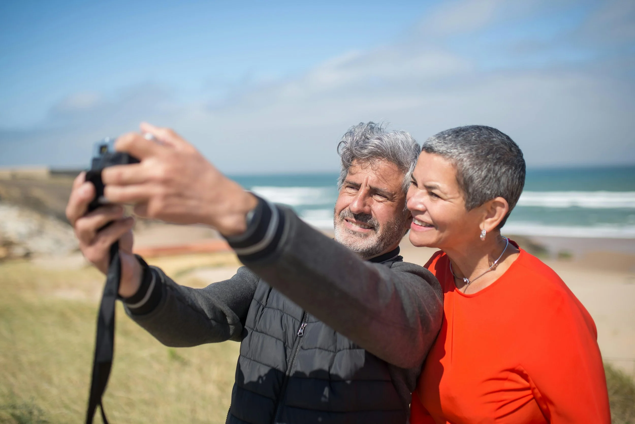 A senior man and woman taking a selfie on a beach with the ocean in the background, both smiling and enjoying a sunny day.