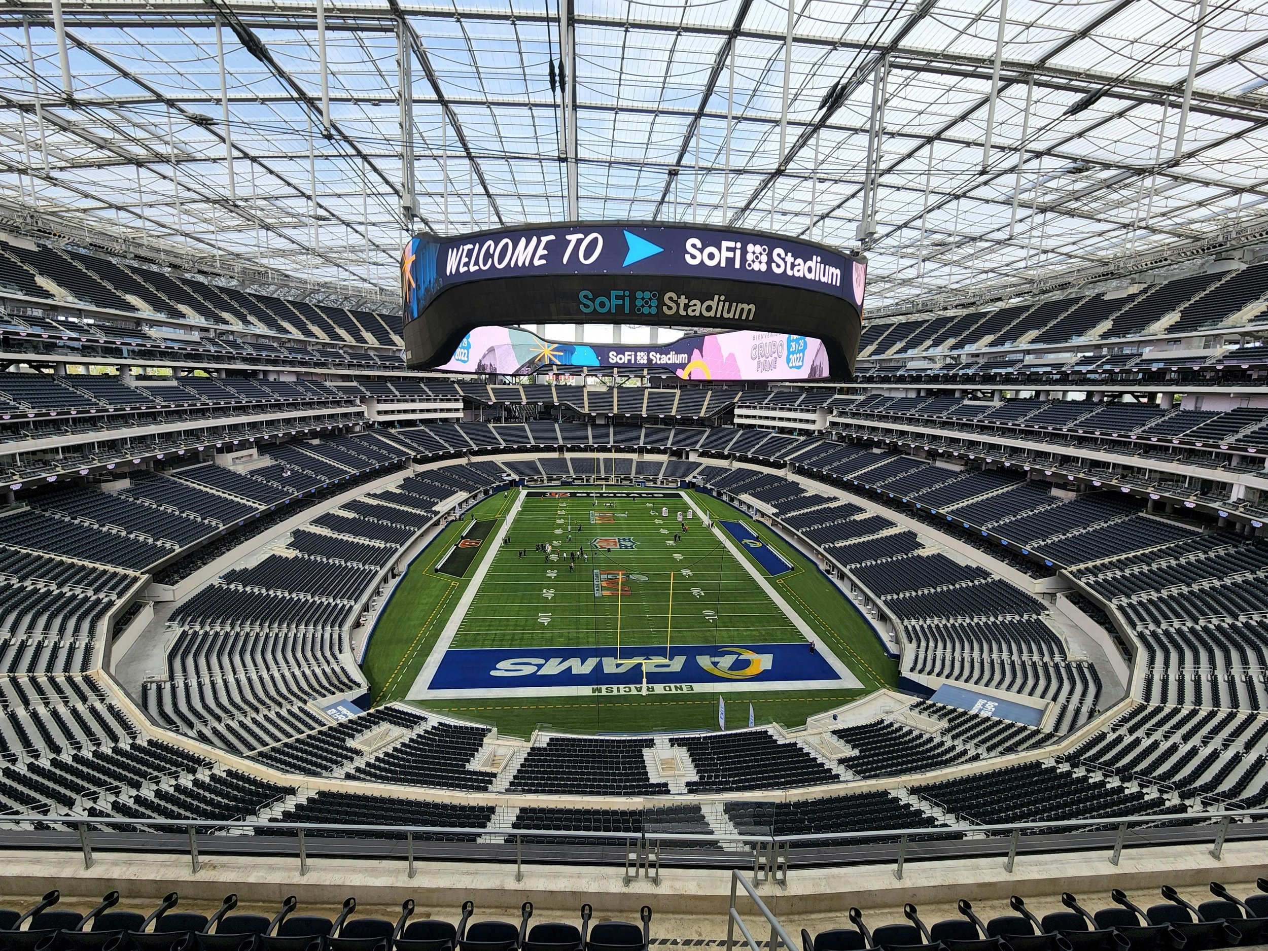 Empty football stadium with a green field, seating all around, and a large scoreboard displaying welcome messages for SoFi Stadium.