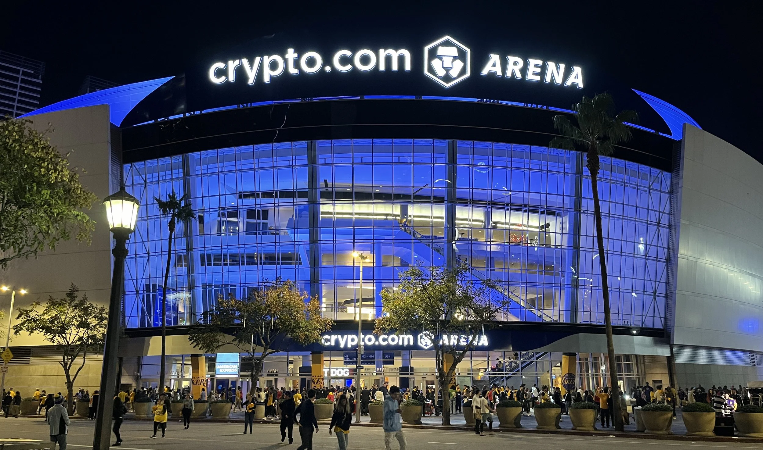 Nighttime exterior view of crypto.com Arena with illuminated signage, glass facade showing interior escalators, and people gathering outside