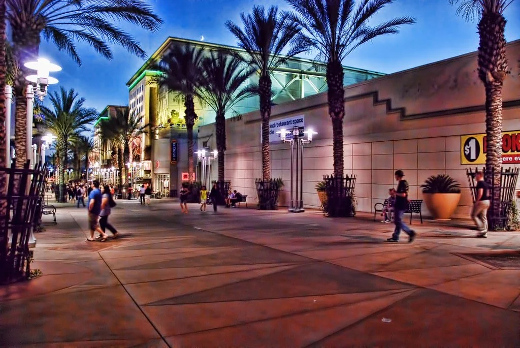 Nighttime scene at a lively outdoor shopping area with palm trees, illuminated buildings, and people walking and sitting on benches.