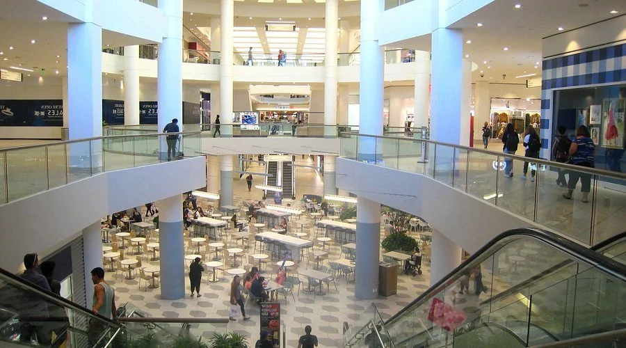 Interior of a multi-level shopping mall with a food court area on the ground floor, people walking and shopping on upper levels, and escalators connecting the floors.