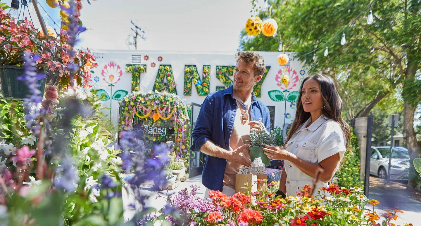 Two people shopping for plants at an outdoor market on a sunny day, surrounded by blooming flowers and greenery, with a decorated white wall in the background.