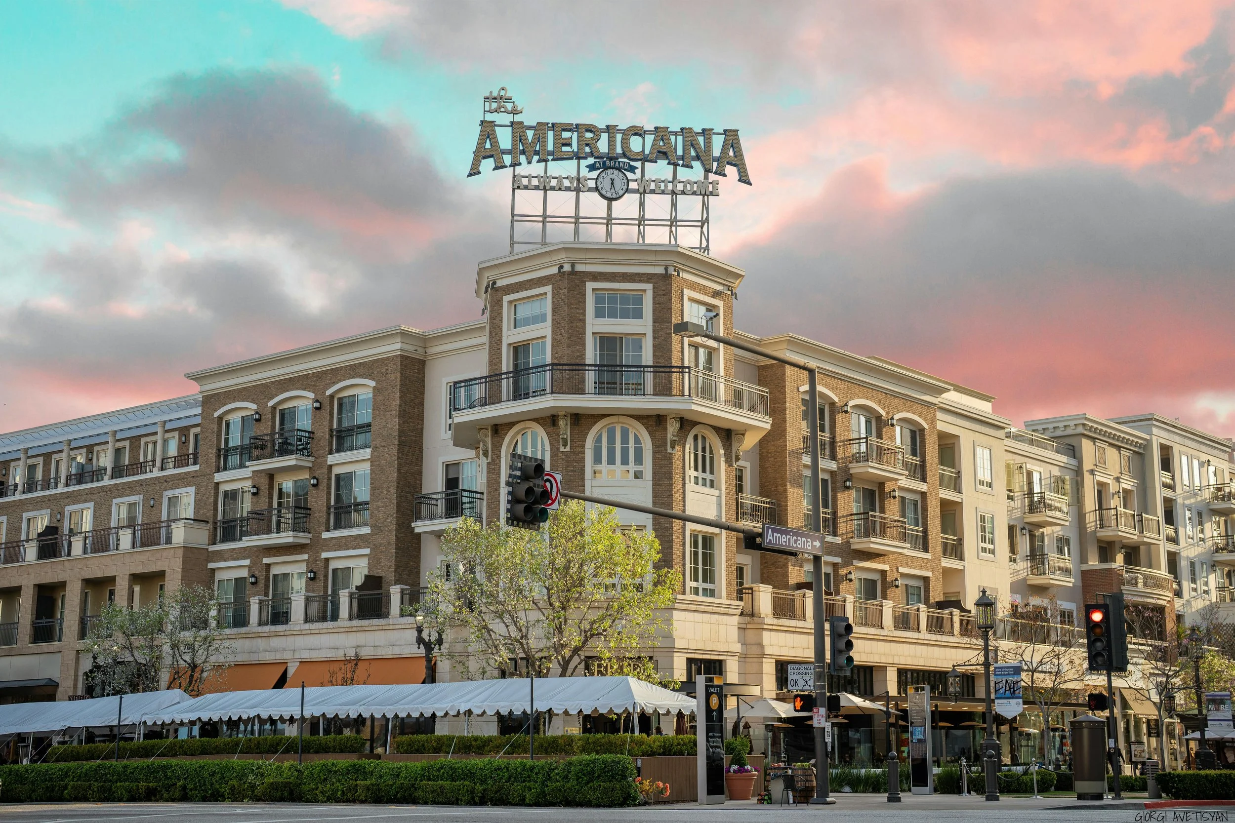 A multi-story building with a sign on top that reads 'The Americana, Always Welcome.' The building has large windows, balconies, and a clock in the sign. The scene is set during sunset with a pink and gray sky. There are trees, a street with traffic signals, and outdoor seating in front of the building.