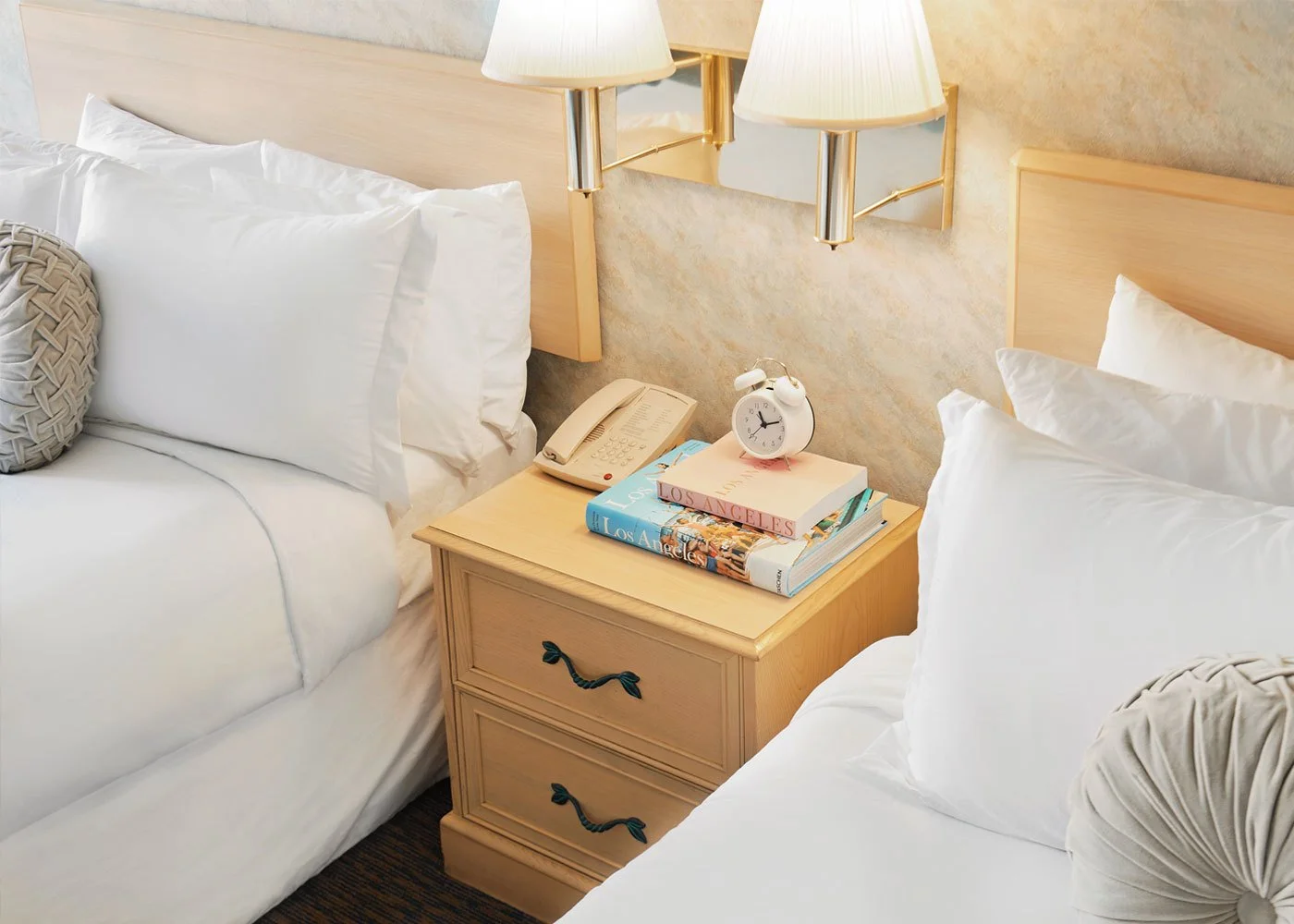 Two beds with white bedding and pillows, a nightstand between them, with a small white alarm clock, a beige telephone, and stacked books titled 'Los Angeles,' a lamp, and a mirror on the wall behind.
