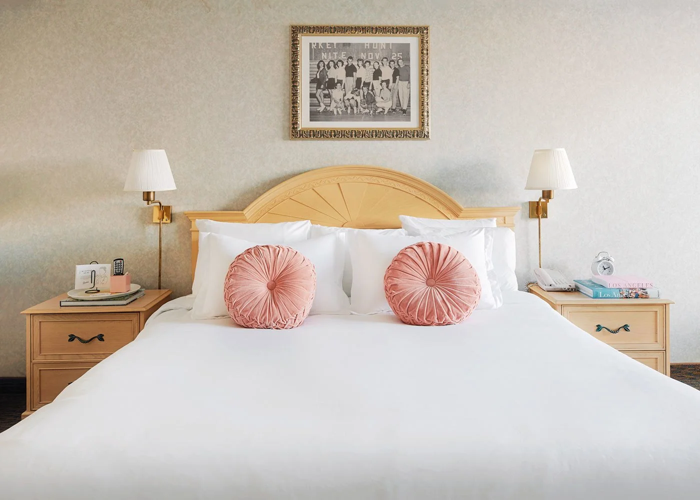 A neatly made hotel bed with white linens, pink round decorative pillows, wooden headboard, and matching wooden nightstands with lamps, books, and a phone in a hotel room.