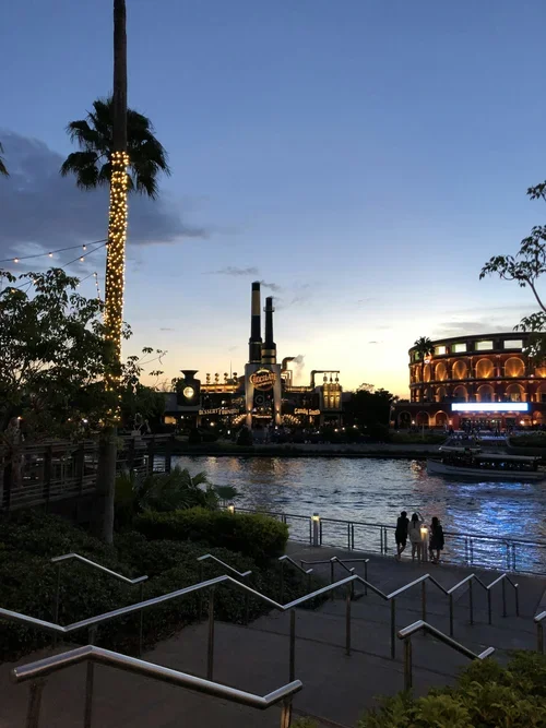 A riverwalk scene during sunset with a lit palm tree, a water body, and illuminated buildings in the background, including a factory and a round hotel, with people walking along the riverbank.