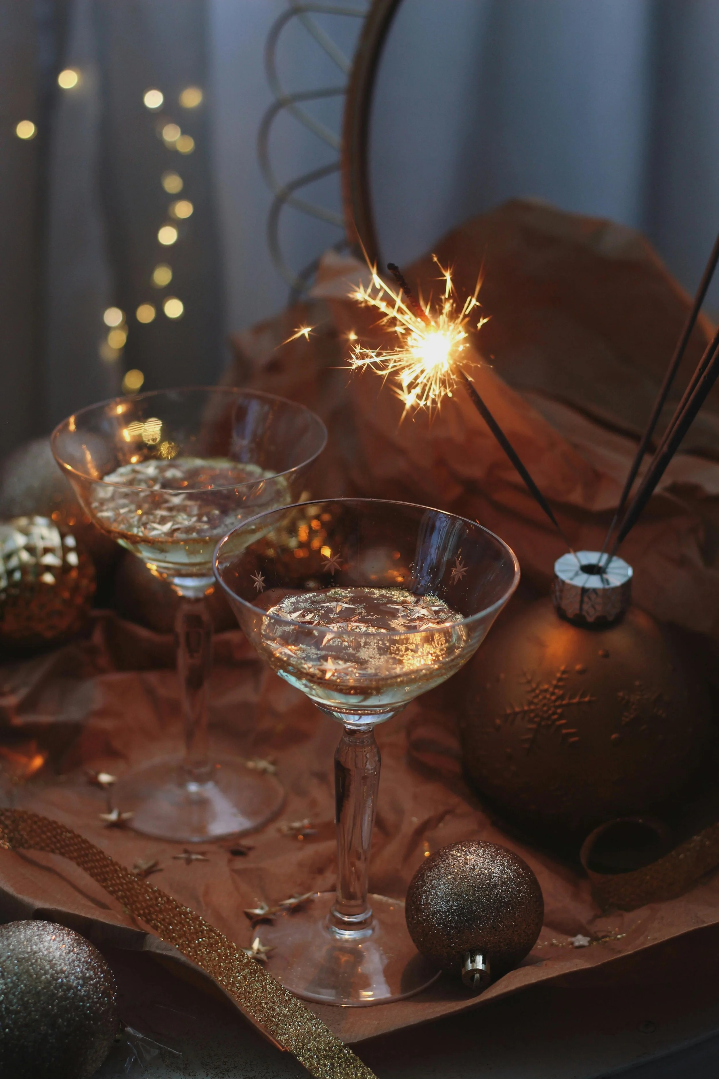 Two martini glasses filled with a sparkling beverage, a lit sparkler, and gold Christmas ornaments on a table with gold ribbon and star decorations, in a festive setting.