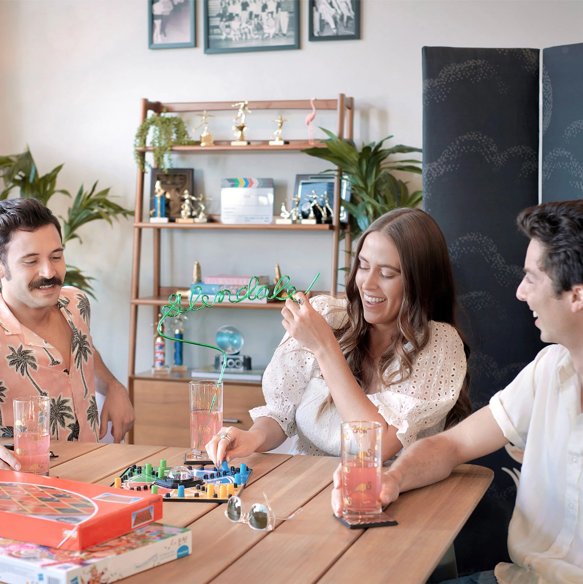 Three people playing a board game at a wooden table, with drinks, in a room with decor including plants, trophies, and framed pictures.