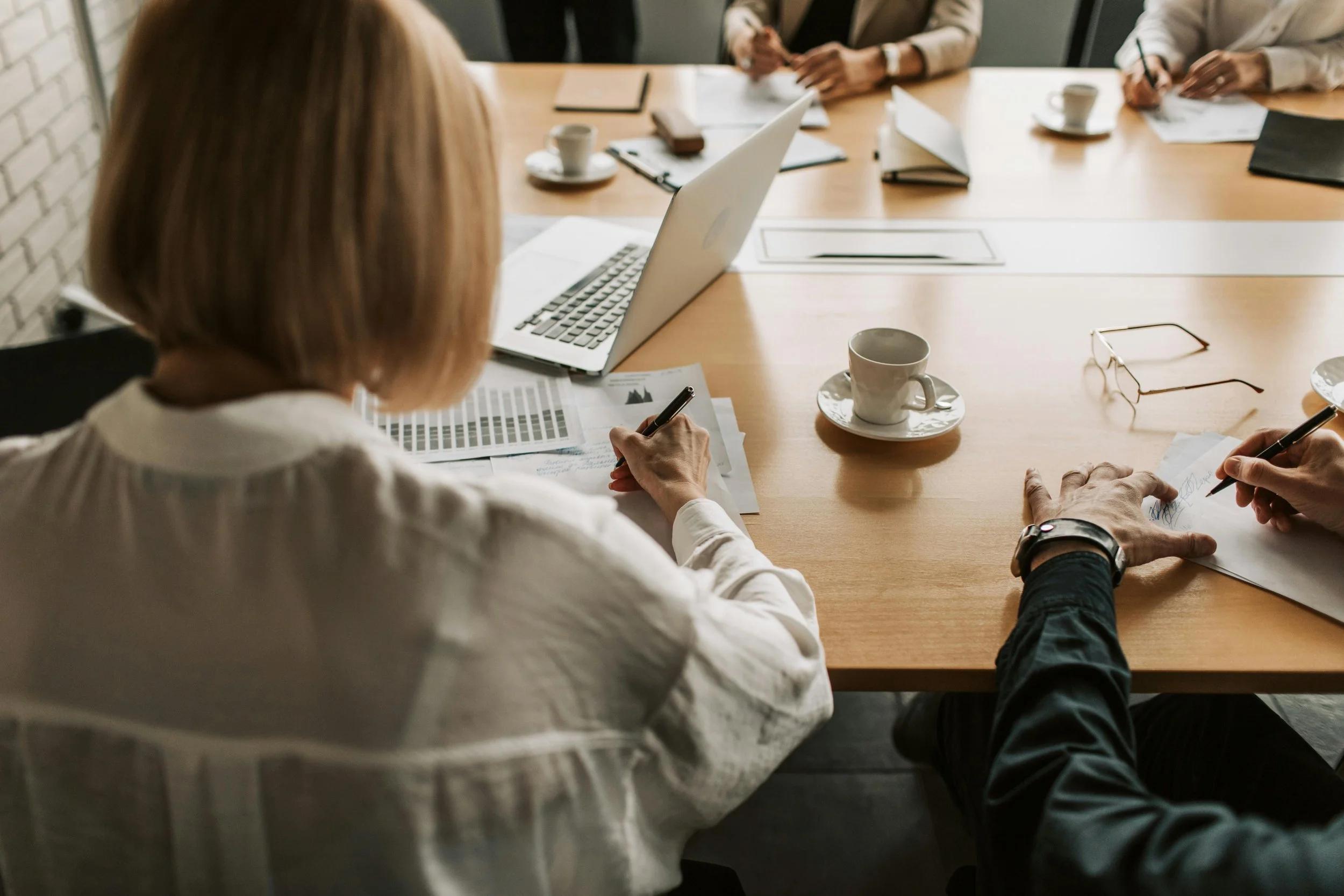 A group of people sitting around a conference table, engaged in a meeting. One person is seen from the back with short blond hair, writing on a paper. There are glasses, cups, a laptop, documents, and notebooks on the table.