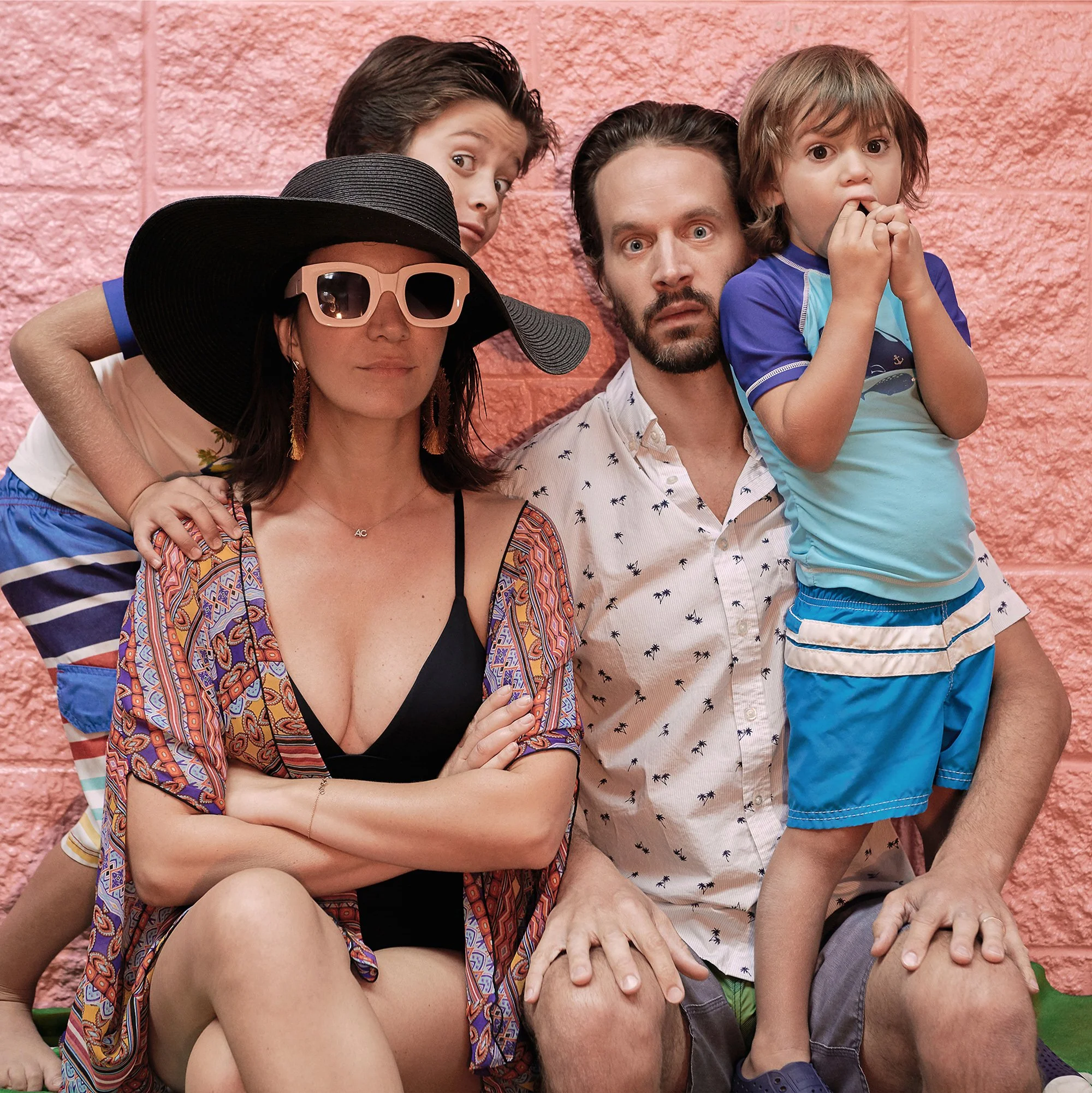 A family of five posing against a pink brick wall. The woman is wearing a large black hat, sunglasses, and a patterned cover-up over a black swimsuit. The man has a beard, is wearing a white shirt with small black patterns. The three children are around them; two boys and a young girl. One boy is leaning over the woman’s shoulder, and the girl is standing on the man’s lap, with all of them looking at the camera with surprised expressions.