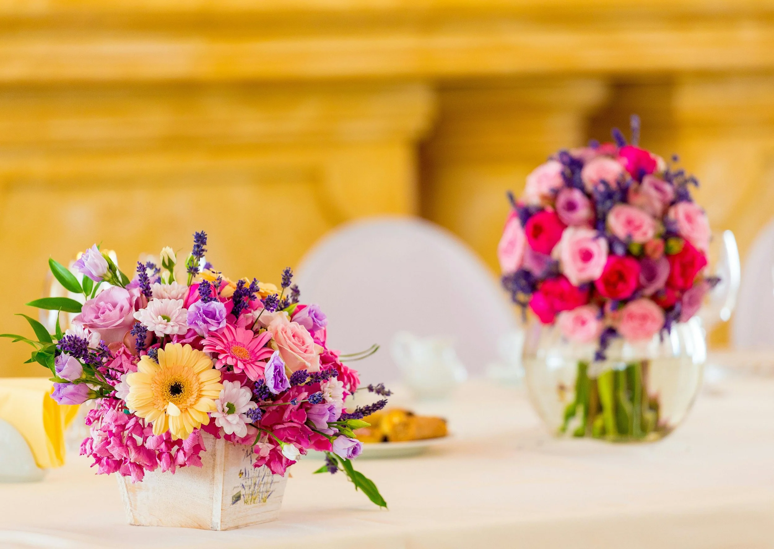 Colorful flower arrangements with pink, purple, yellow, and red flowers on a table, with blurred chairs and background.