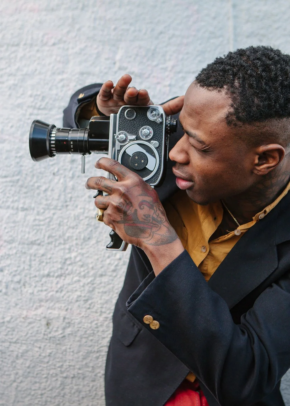 A tattooed man, dressed in a black blazer and tan shirt, is looking through a vintage film camera mounted with a lens, against a light gray textured wall.