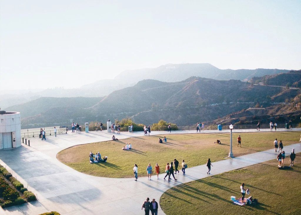 People walk, sit, and gather on a rooftop with a grassy area, surrounded by mountain scenery.