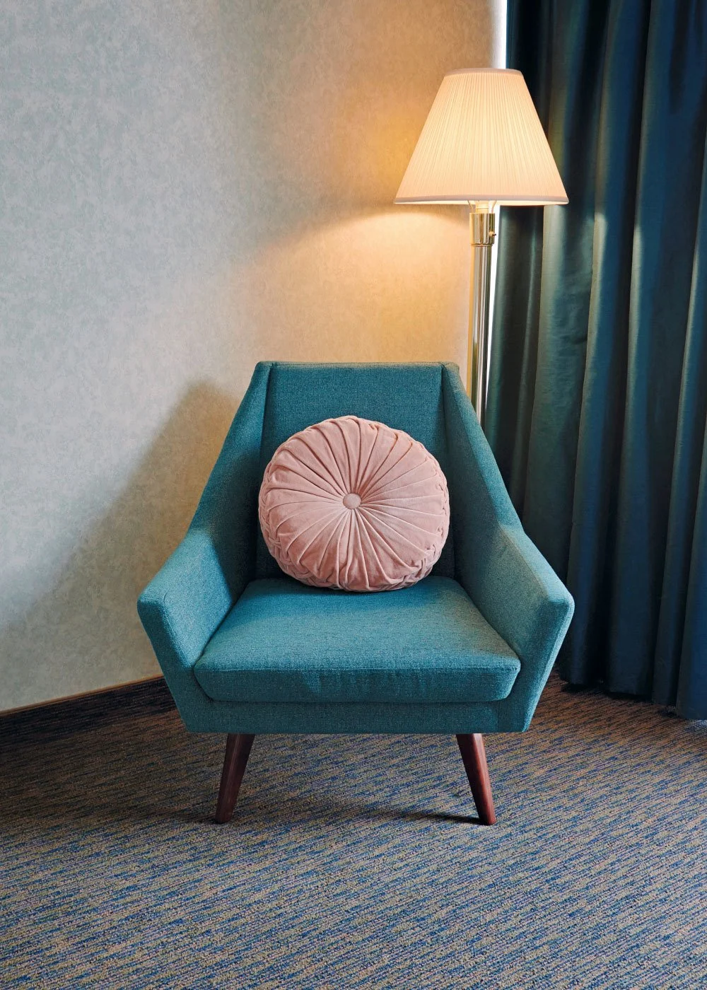 A teal armchair with wooden legs and a round pink cushion, placed beside a standing lamp with a white shade in a room with beige textured walls and blue curtains.