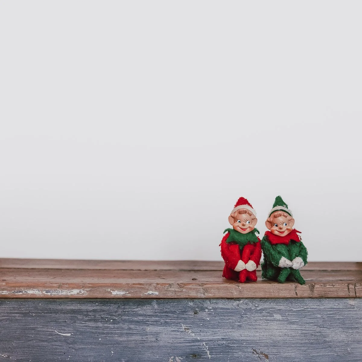 Two small elf dolls with cheerful faces, one dressed in red and the other in green, sitting on a rustic wooden surface against a plain white background.