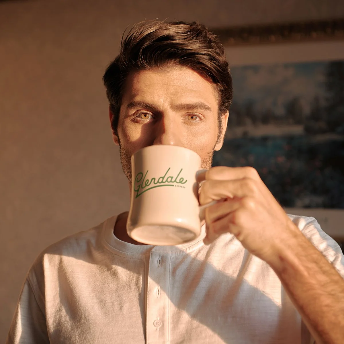 A man with brown hair and blue eyes drinking from a latte mug labeled 'Glendale' in a warmly lit room.
