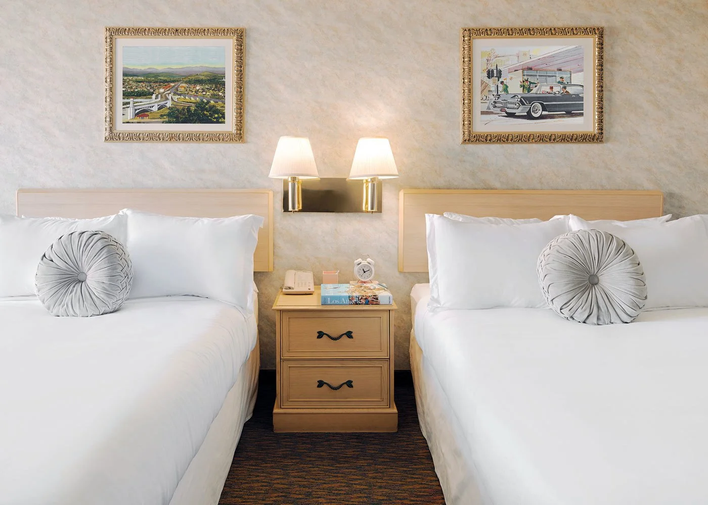 Hotel room with two beds, bedside table, wall-mounted lamps, and framed artwork, including a landscape and a vintage car scene.