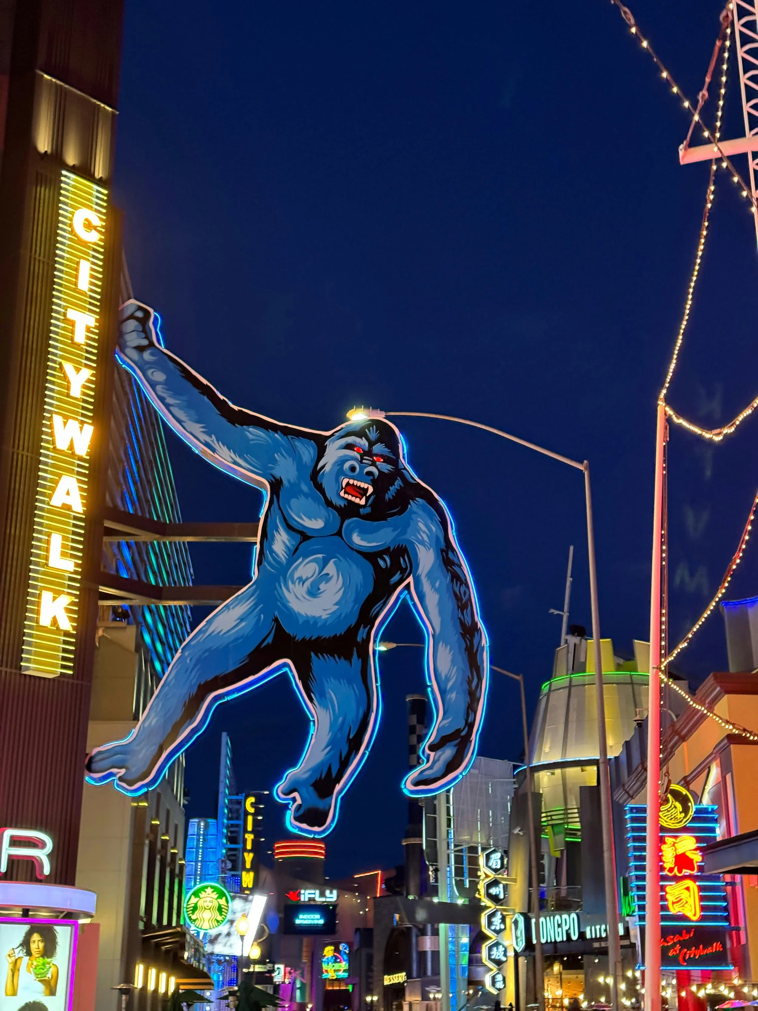 Neon sign of a giant, blue, cartoon-like Gorilla hanging from a pole at night in a busy city street with colorful lights and signs.