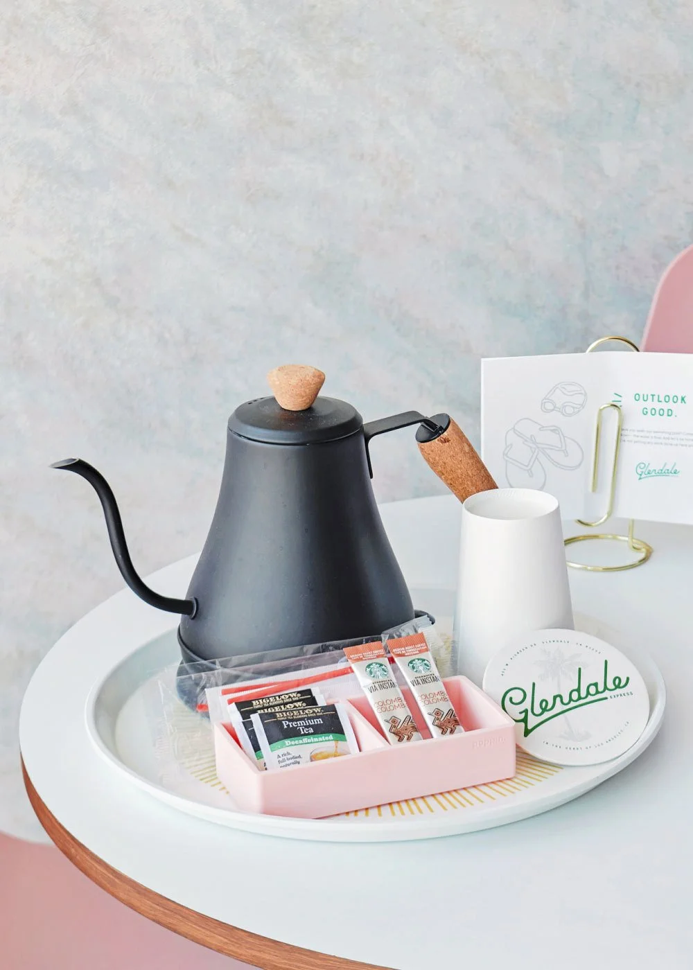 Black gooseneck electric kettle with cork handle on a white table, next to a tea set and white Glendale coaster and cup..
