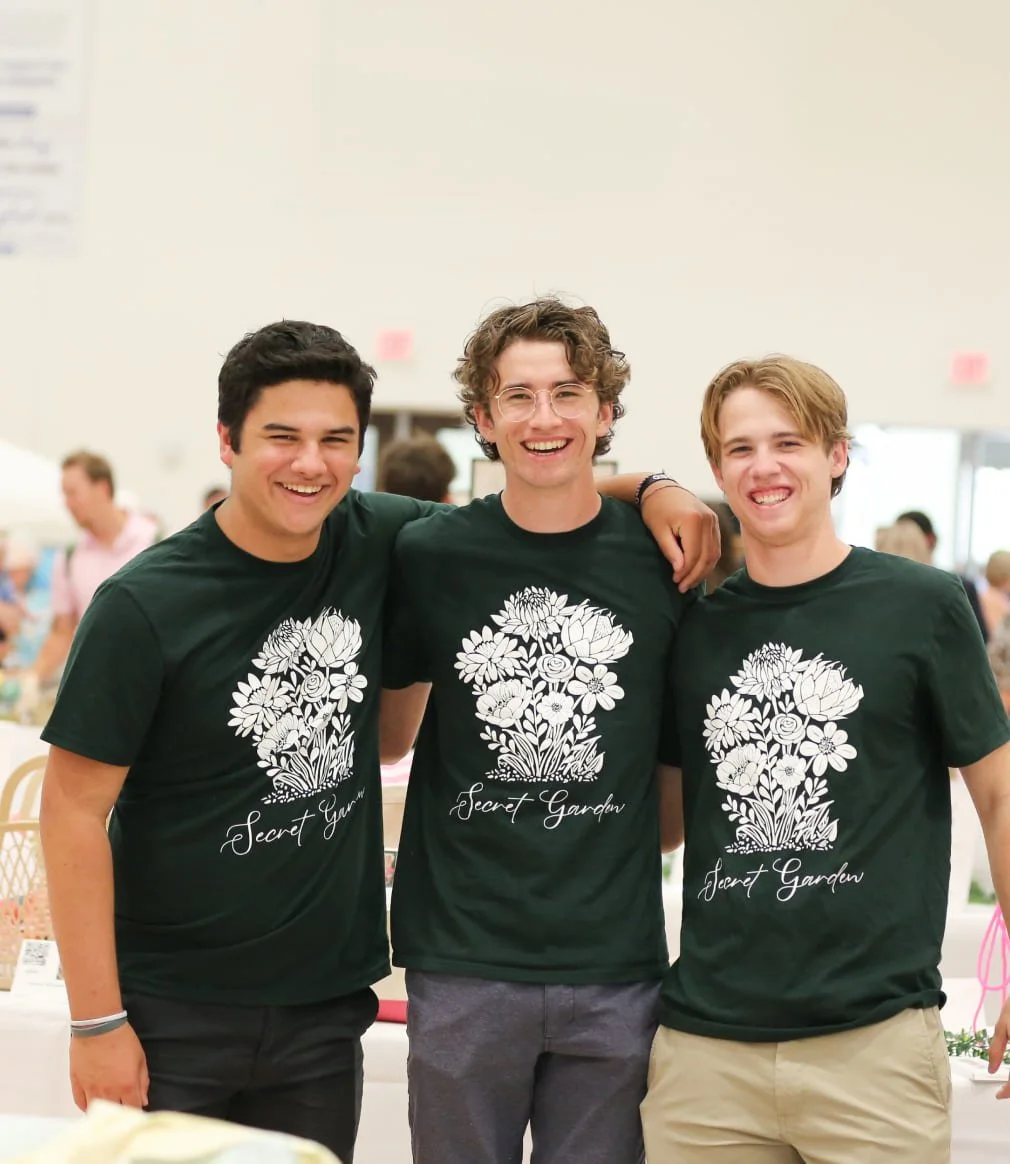 Three young men smiling and standing together in a bright indoor space, wearing matching black t-shirts with white floral designs and the words 'Secret Garden' printed on them.