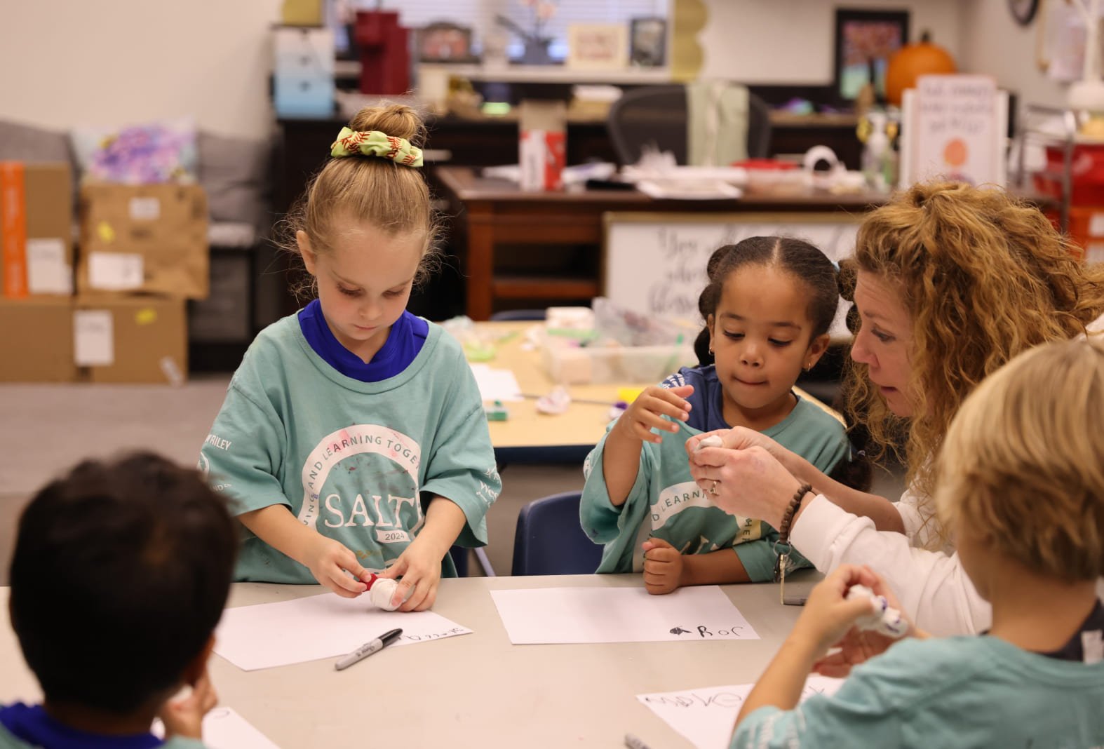 A teacher assists a young girl with a craft project at a table with children participating in a classroom activity.