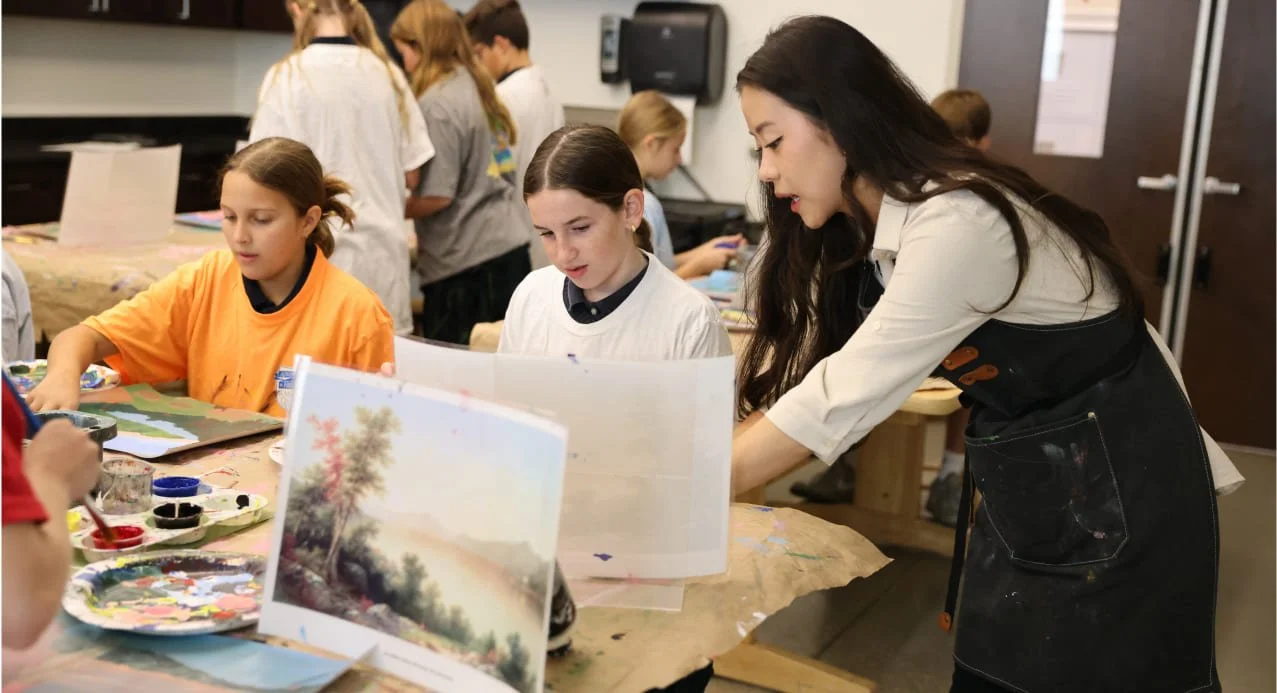 Teacher helping students with an art project in a classroom, painting supplies on the table.