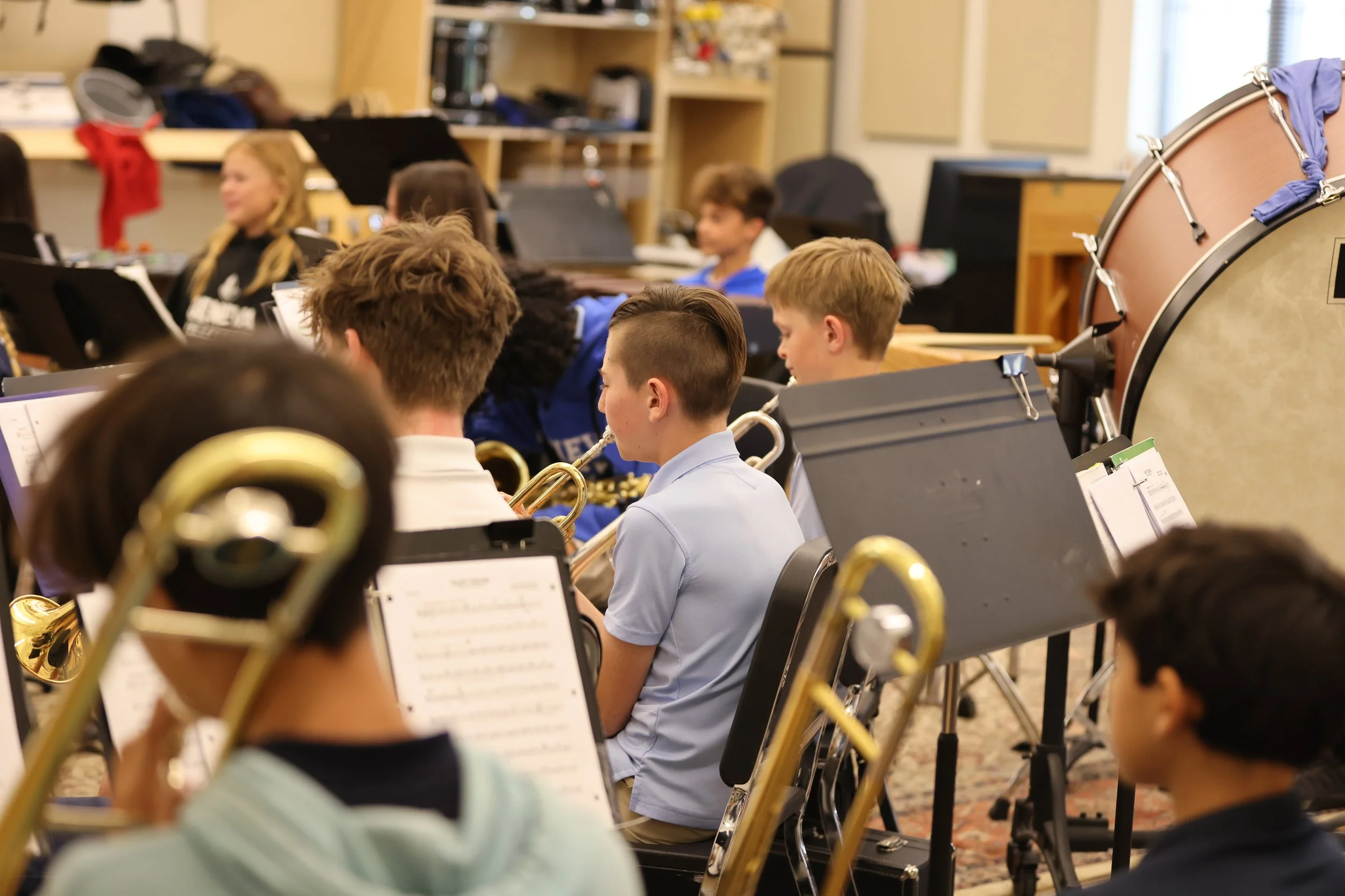 Students in a classroom playing musical instruments, with a focus on brass instruments and a large bass drum to the right.