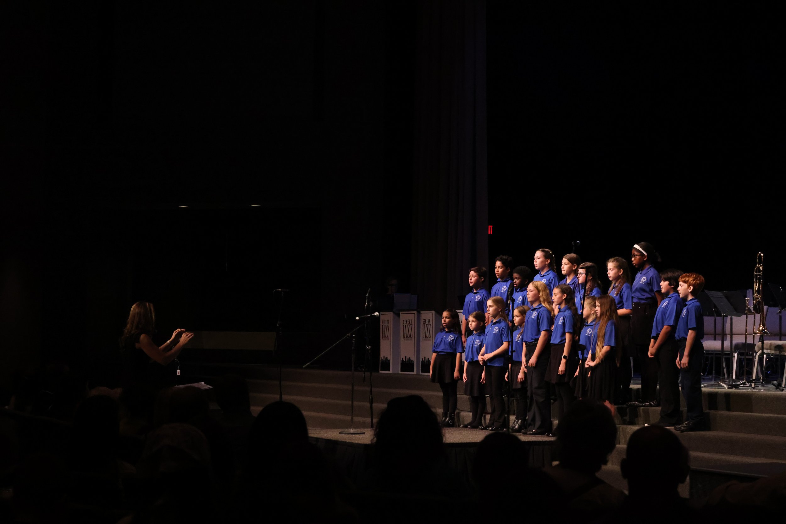 A group of children in blue uniforms singing on stage with a music stand and a saxophone, while a conductor directs them in a dark auditorium.