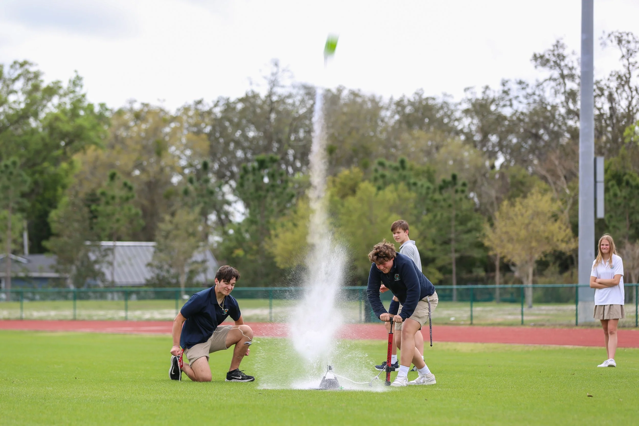 Group of students standing on a grassy field during daytime, with one student launching a model rocket and others observing near a firefighting foam.