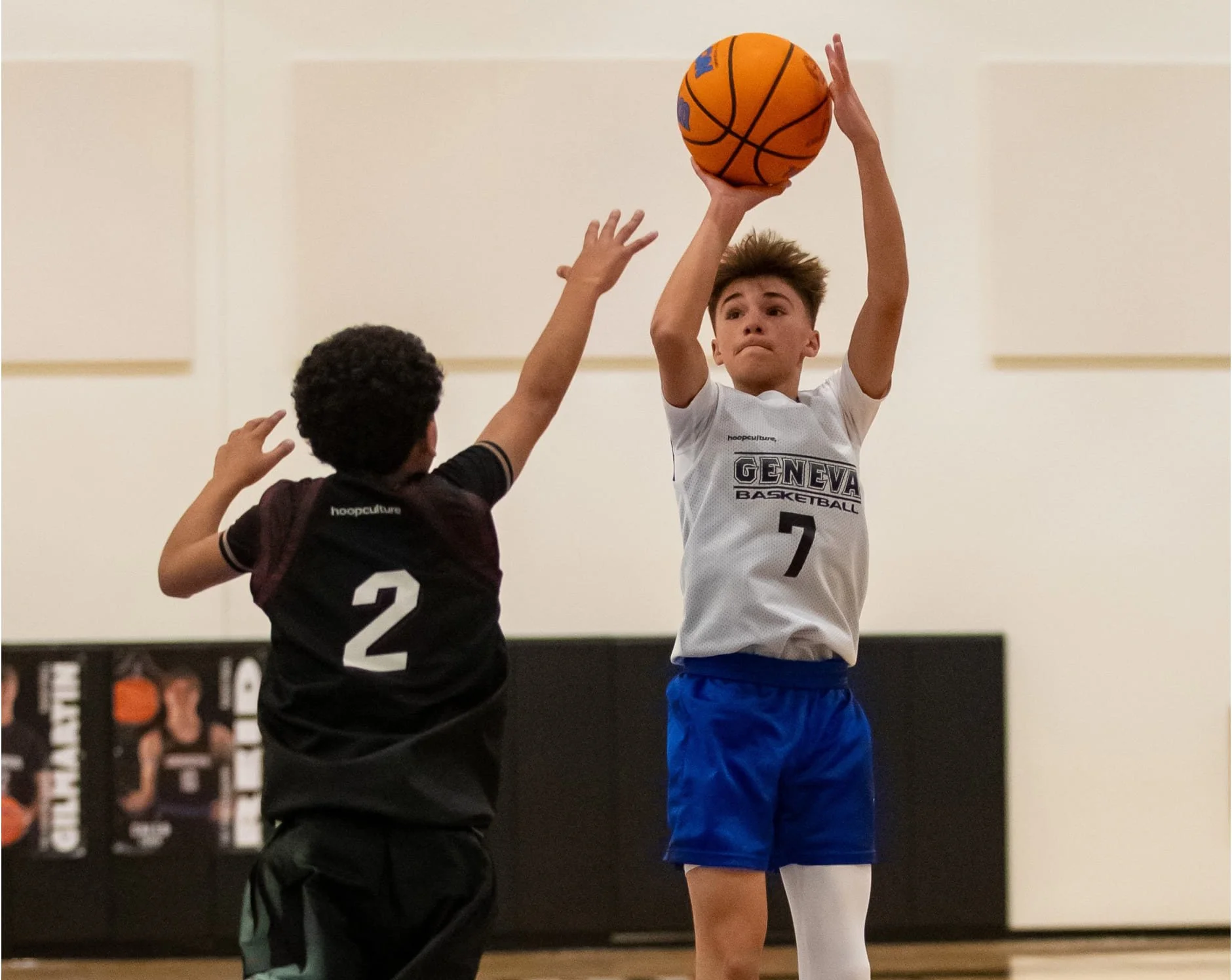 Two young male basketball players, one in a white jersey holding a basketball and jumping, the other in a black jersey defending, indoors on a basketball court.
