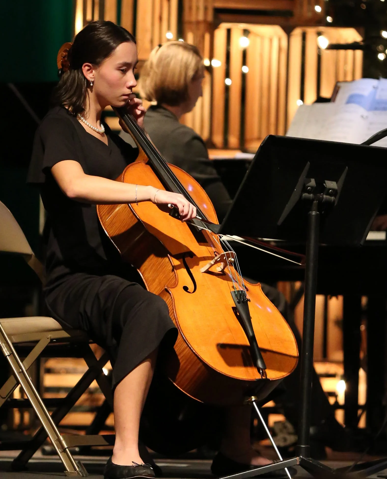 Student in black attire playing a cello during a musical performance in a dimly lit studio with wooden panels and sheet music on a stand.