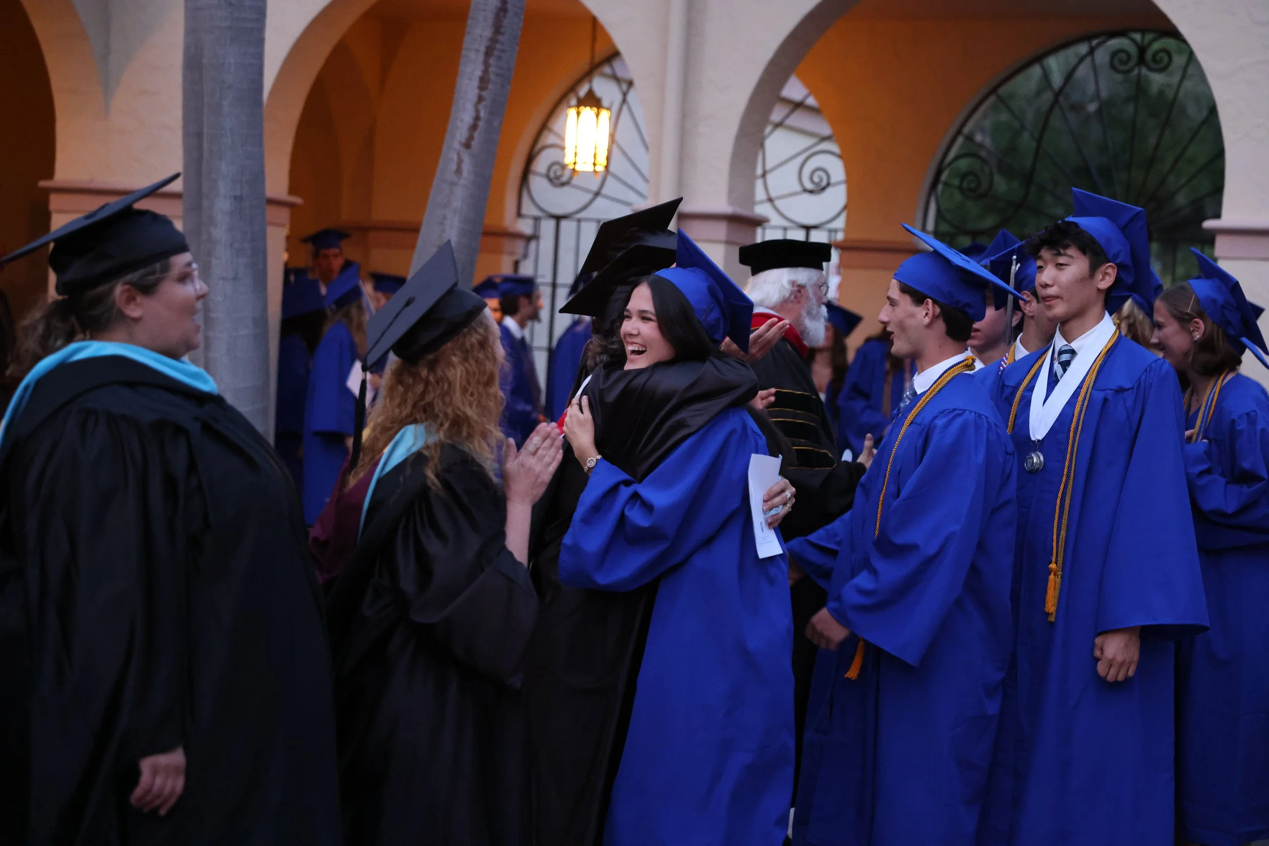 Graduate students in caps and gowns at a commencement ceremony, shaking hands and congratulating each other indoors with arches and a warm lighting backdrop.