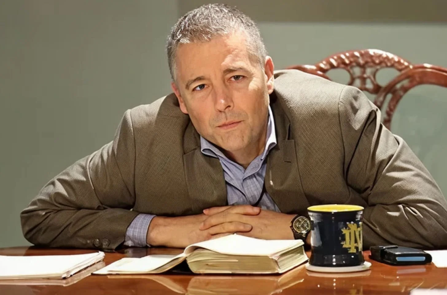A man in a suit leans on a wooden desk with open notebooks, a coffee mug, and a voice recorder, looking tired or bored, in an office setting.