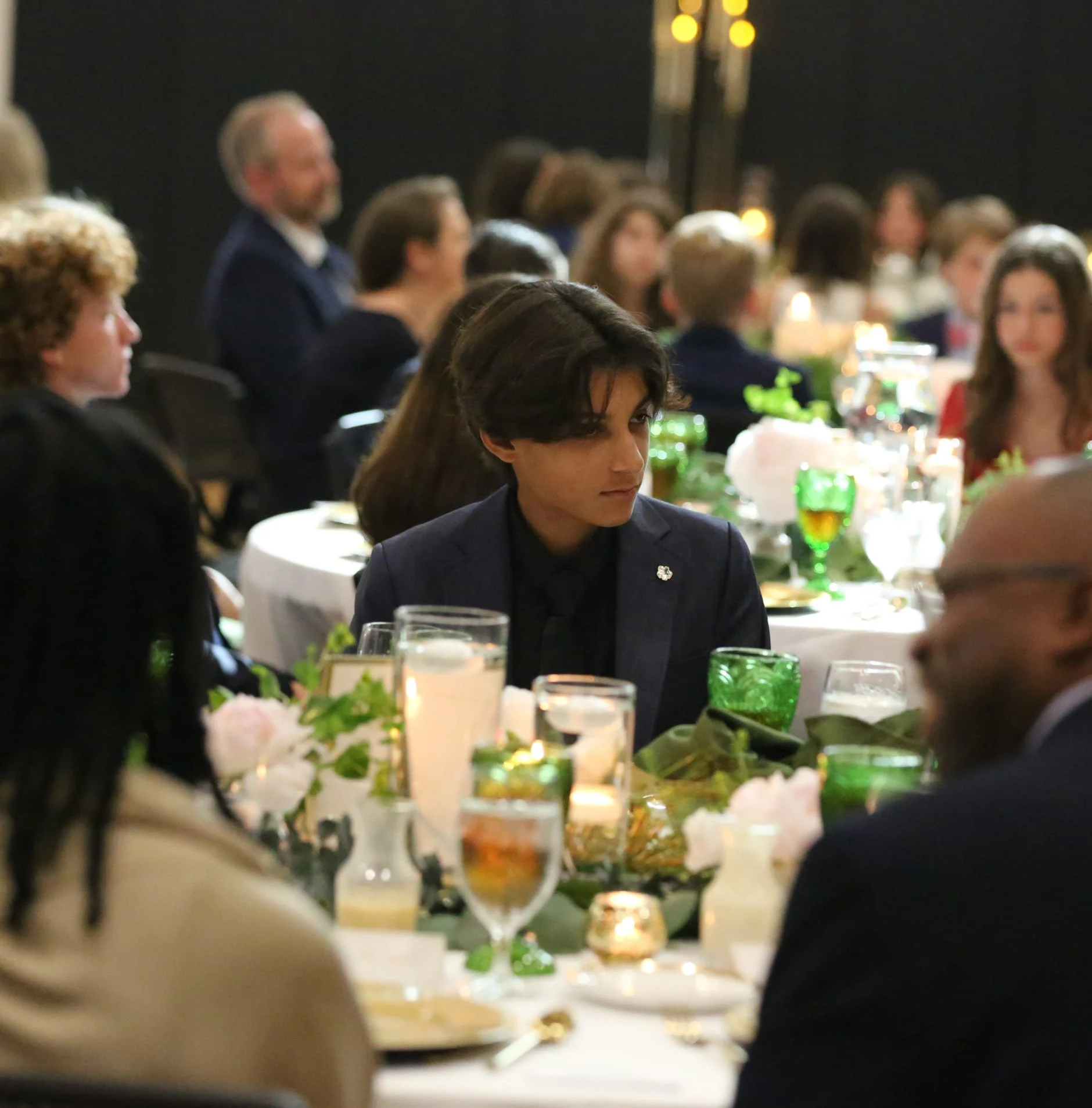 People seated at a formal dinner event, with table settings, candles, and floral arrangements, in a dimly lit setting.