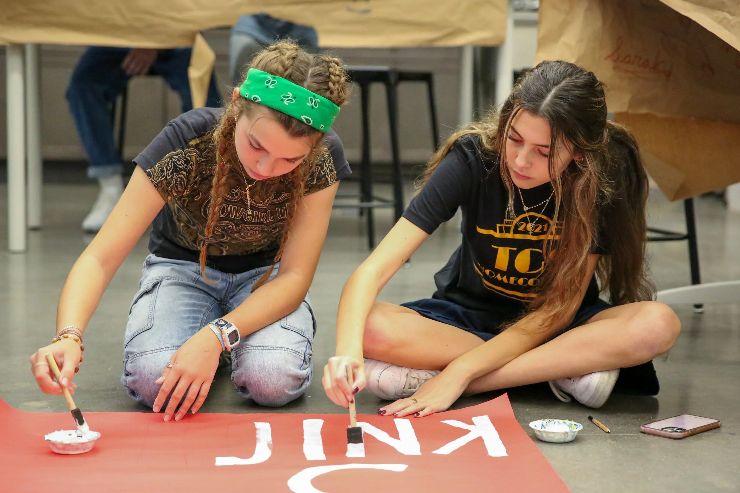 Two young women painting letters on a large red banner on the floor. They are sitting cross-legged, focused on their work, with bowls of white paint and brushes nearby.