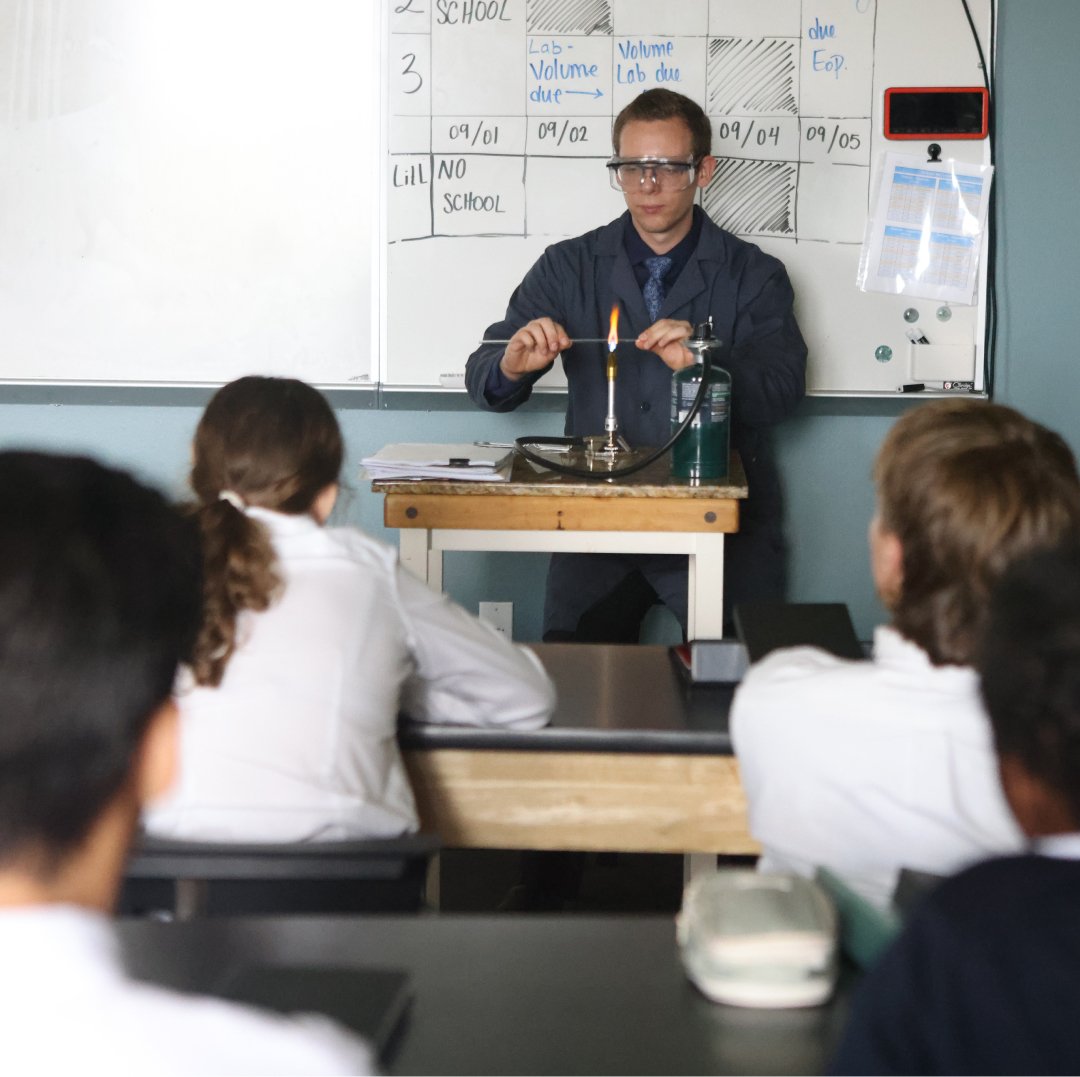 A science teacher demonstrates a chemistry experiment with a lit flame in a classroom, with students observing at their desks.