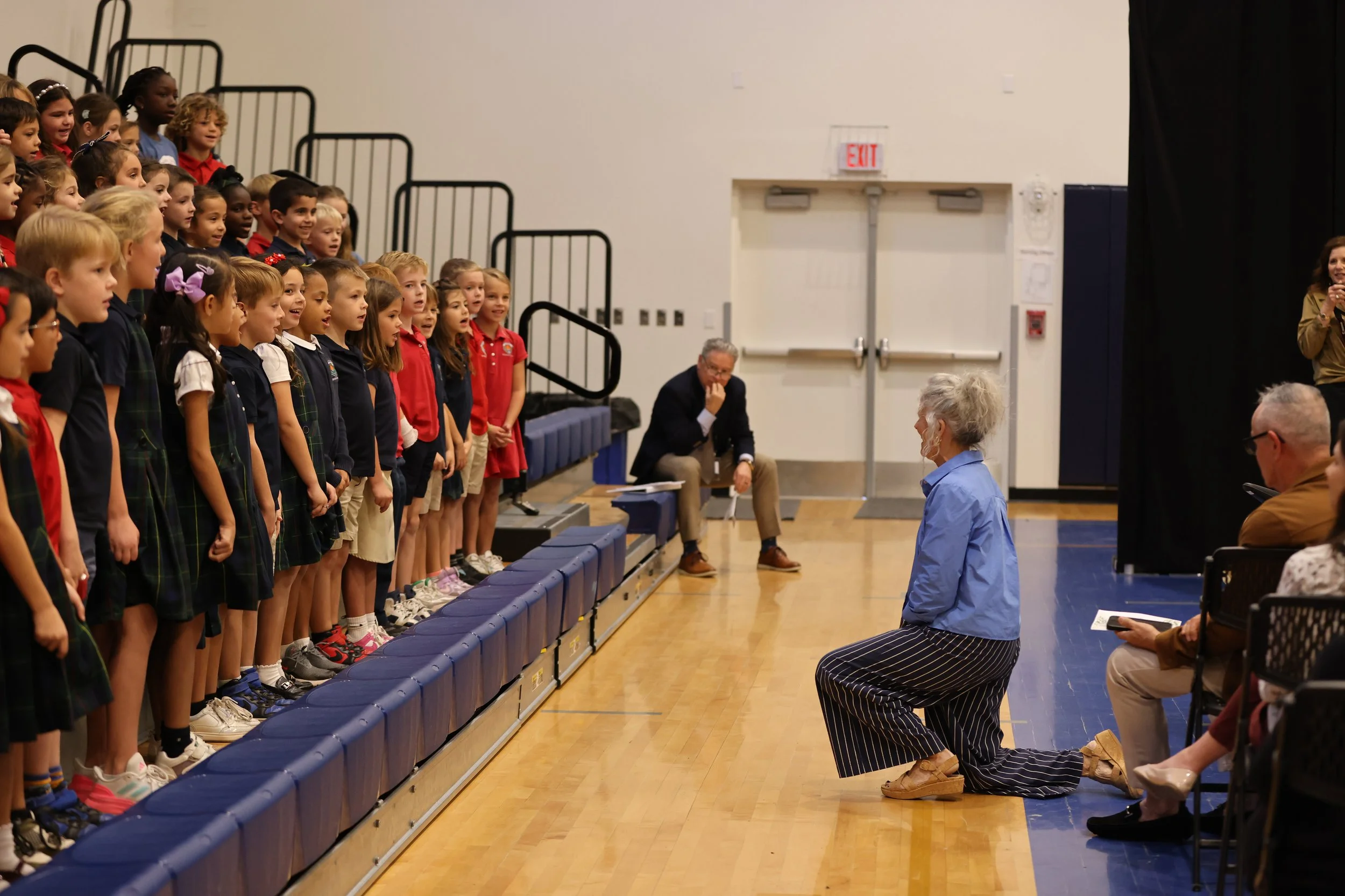 A choir of young children standing on risers, singing, directed by a conductor kneeling in front.